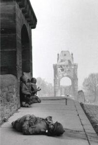 Black-and-white wartime photograph, circa March 26, 1945, showing PFC Charles Theodore Plog lying on the Ernst Ludwig Bridge (now the Nibelungen Bridge) in Worms, Germany, while two U.S. Army servicemen take cover near the bridge's western tower. The bridge's damaged eastern tower is visible in the background.